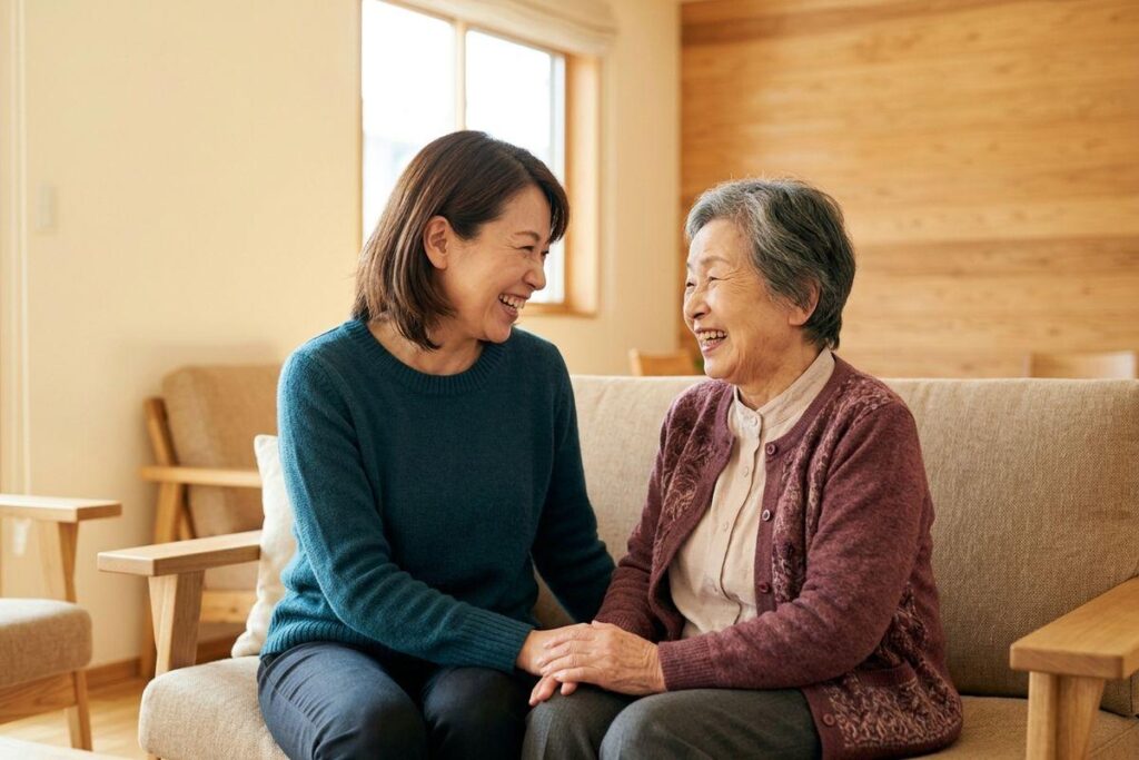 A younger woman and a senior sit on a sofa, holding hands and smiling at each other.